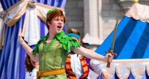Person dressed as Peter Pan, wearing a green costume with a green hat, performing energetically on stage. Holding a sword in one hand and displaying a determined expression, they stand against a backdrop of blue and white curtains and a stone structure.