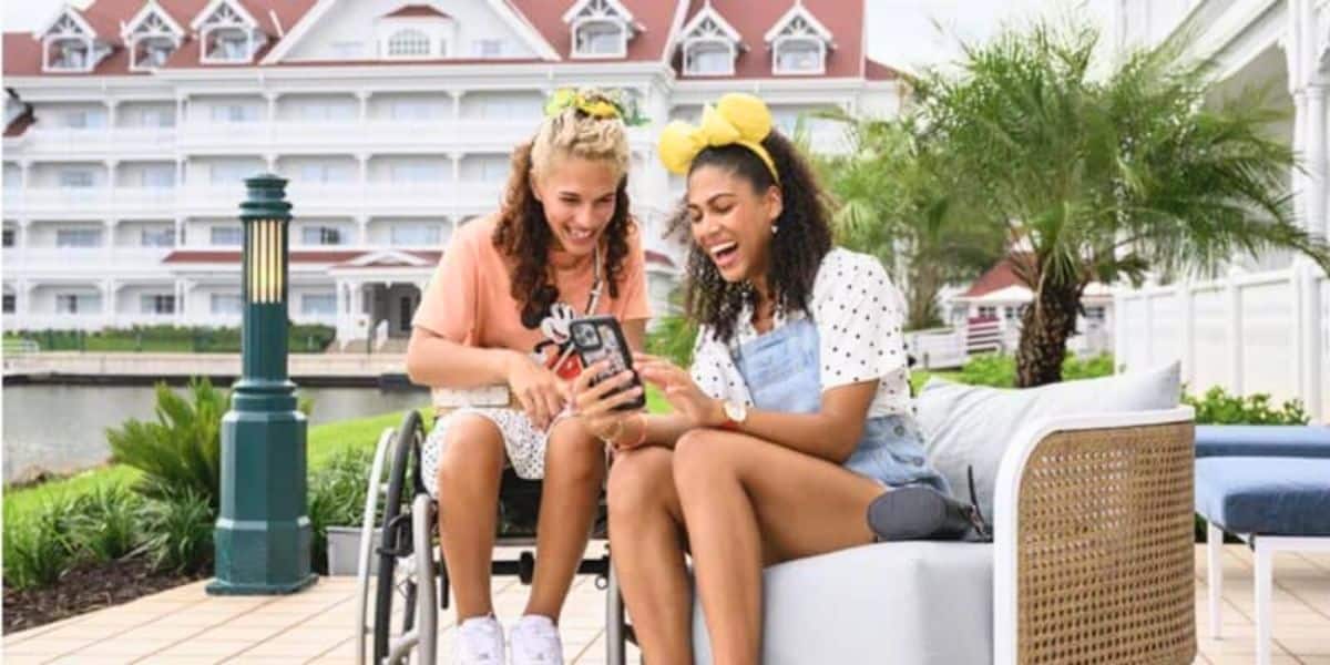 Two women are sitting outside a building with red roofs. One is in a wheelchair, wearing a peach-colored shirt, and the other is on a bench, wearing a white shirt and denim overalls with yellow Minnie Mouse ears. Both are smiling and looking at a smartphone.