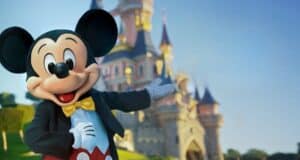 Mickey Mouse, dressed in his classic red shorts, yellow bow tie, and black jacket, stands with one arm outstretched in front of a fairytale castle at Disneyland. The sky is clear and blue, creating a magical, inviting atmosphere.