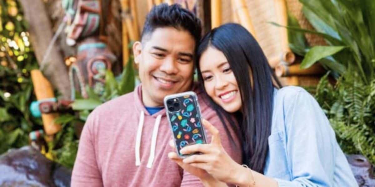 A happy couple takes a selfie together with a smartphone, smiling at the camera. The man wears a red hoodie, and the woman wears a blue shirt. They are outdoors, surrounded by lush greenery and colorful, decorated wooden pillars in the background.