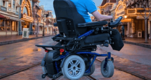 A person wearing a blue shirt is seen from behind using a motorized wheelchair on an empty street lined with buildings adorned with lights, reminiscent of the enchanting ambiance created for Disney guests. The atmosphere suggests a festive or amusement park setting.