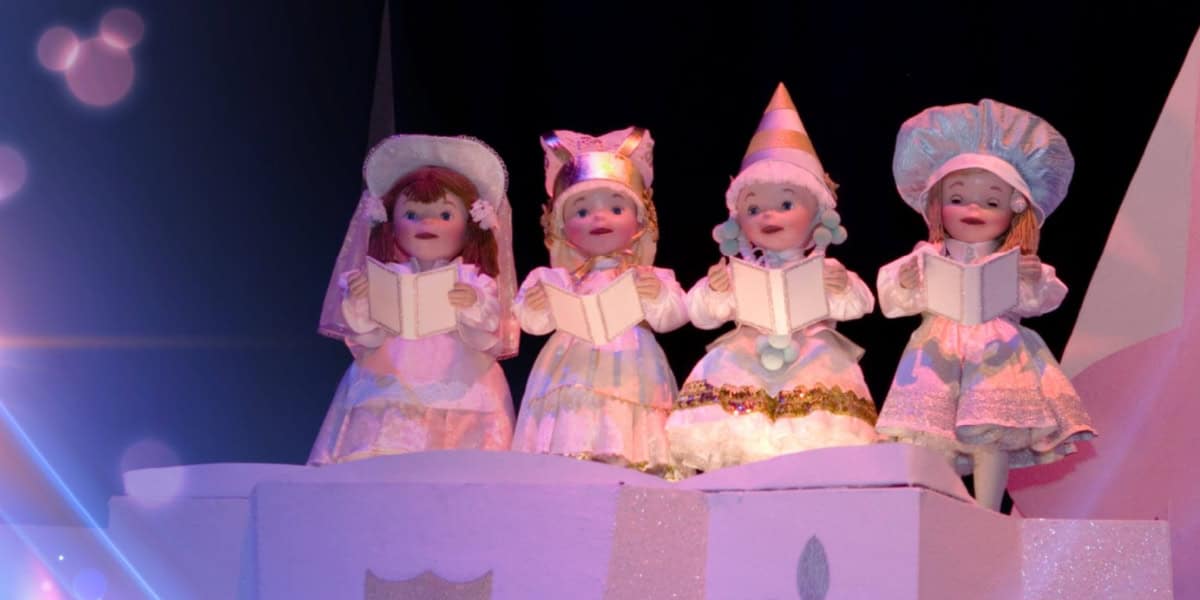 Four childlike dolls dressed in whimsical, old-fashioned costumes stand in a row, each holding an open book as if singing. The background is dark with a bluish lens flare effect on the left, adding a magical ambiance to the scene.