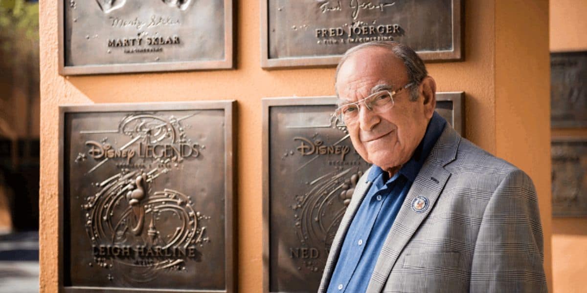 An older man with short hair and glasses stands in front of several plaques on an outdoor wall. Wearing a light gray blazer over a blue shirt, he looks at the camera. The plaques feature the names and signatures of various Disney Legends honorees, perhaps inspiring this Disney Imagineer.
