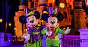 Minnie and Mickey Mouse are dressed in bright green Halloween costumes with a castle illuminated in orange behind them. Minnie wears a witch outfit, and Mickey sports a top hat and bow tie. They stand on a bridge decorated with lights at night, posing for a photo.