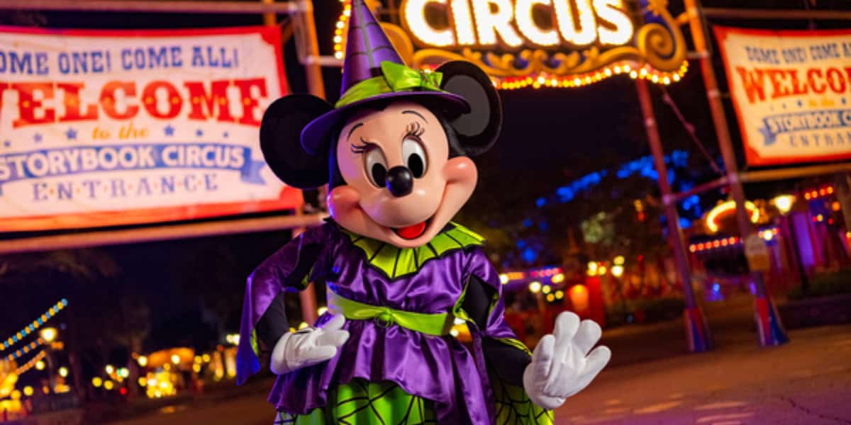 Minnie Mouse is dressed in a vibrant purple and green witch costume with an orange bow, smiling and posing in front of a brightly lit circus-themed background with banners reading "Welcome to the Storybook Circus." The scene is colorful and festive.