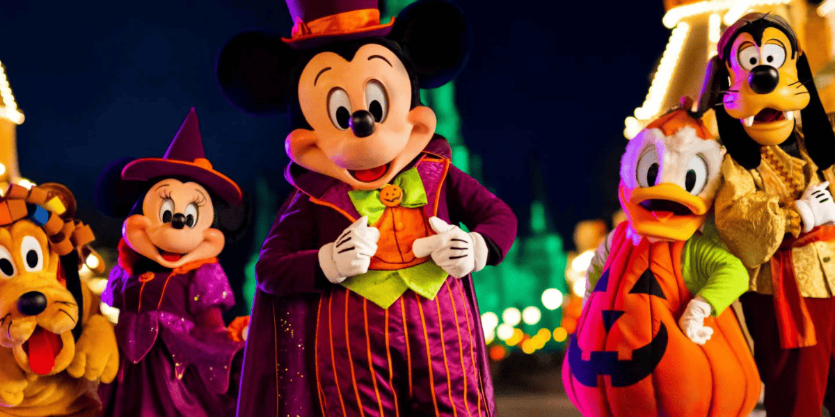 Mickey Mouse and friends, dressed in Halloween costumes, pose for a photo in front of a brightly lit castle. Mickey wears a magician's outfit, Minnie is a witch, Donald Duck is a pumpkin, Goofy is a scary character, and Pluto has a festive collar.