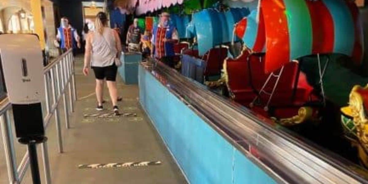 A few people stand in line at an indoor amusement park ride inspired by Peter Pan. The ride vehicles are colorful and whimsical with an overhead canopy. Social distancing markers are on the ground, and a hand sanitizer station is visible on the left.