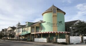Colorful building with a pastel green facade, red and white striped awnings, and a cylindrical tower attached. A sign reads "Disney's Saratoga Springs Resort & Spa," one of the renowned Disney Resorts. Several bicycles are parked nearby, and the sky is partly cloudy.