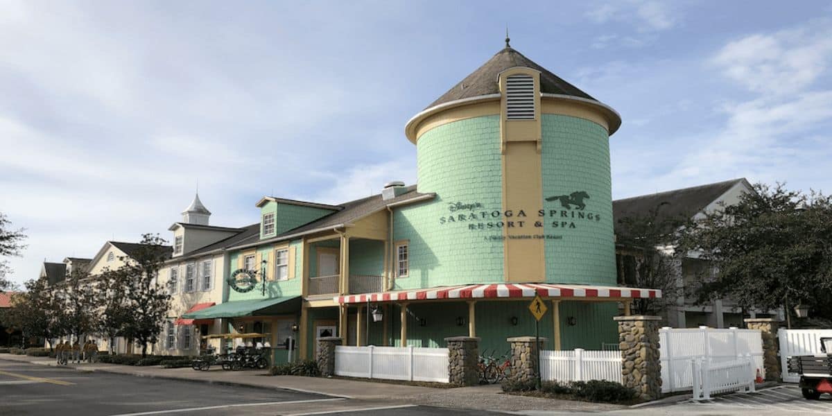 Colorful building with a pastel green facade, red and white striped awnings, and a cylindrical tower attached. A sign reads "Disney's Saratoga Springs Resort & Spa," one of the renowned Disney Resorts. Several bicycles are parked nearby, and the sky is partly cloudy.