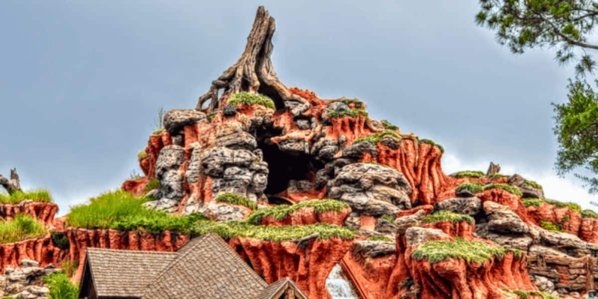 A colorful and detailed rock formation resembling a mountain, with rugged terrain, patches of greenery, and a distinct peak. The sky is overcast, adding contrast to the vibrant hues of the rocks. At its base, a wooden structure with a peaked roof hints at the Magic Kingdom's newest attraction launching next month.