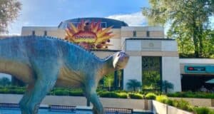 A statue of a dinosaur stands proudly in front of a building adorned with a "Dinosaur" sign and an eye-catching colorful explosion graphic. The scene, reminiscent of Disney's artistry, is framed by lush trees while visitors gather near the entrance.