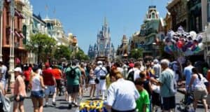A bustling crowd walks down a vibrant street at a theme park, lined with colorful buildings and ornate shops. Balloons of various shapes and colors float on the right side. In the distance, a majestic castle stands tall under a clear blue sky.