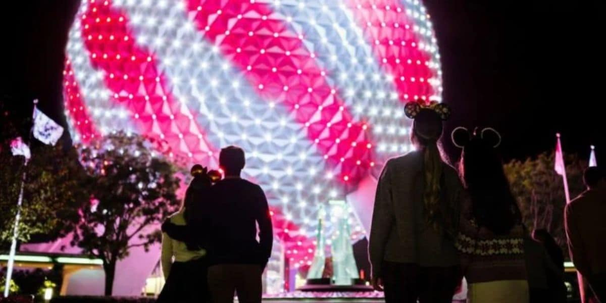 A nighttime scene at Epcot with a family of four wearing Mickey Mouse ears admiring the large, illuminated Spaceship Earth. The geodesic sphere is lit up with bright, colorful patterns, primarily in red and white, creating a festive atmosphere.