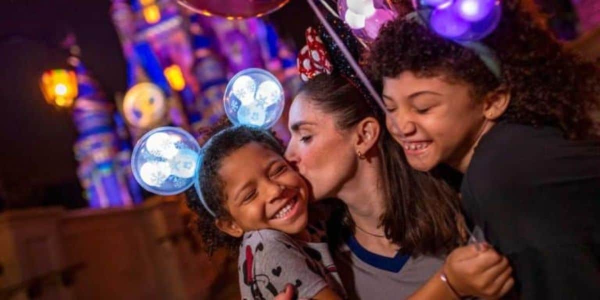 A joyful mother kisses one of her two smiling children, both wearing Mickey Mouse ear headbands, in front of a colorful, illuminated castle at night during Disney After Hours.