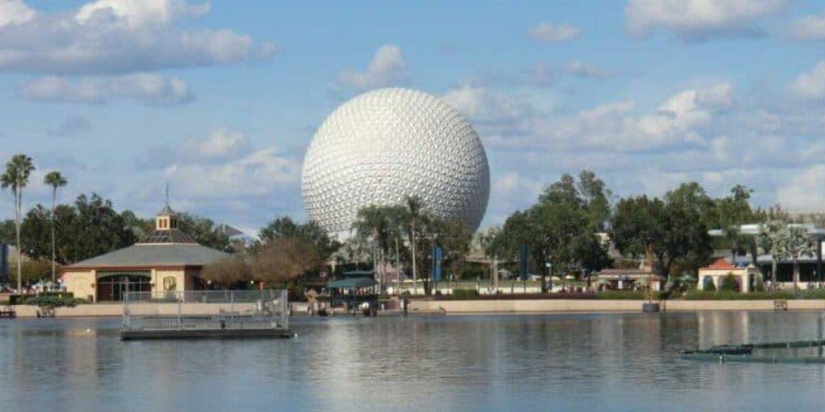 A large white geodesic sphere, known as Spaceship Earth, stands prominently in the background at Epcot theme park in Disney World. The foreground features a calm body of water reflecting the sphere, with buildings and trees on the shoreline under a partly cloudy sky.