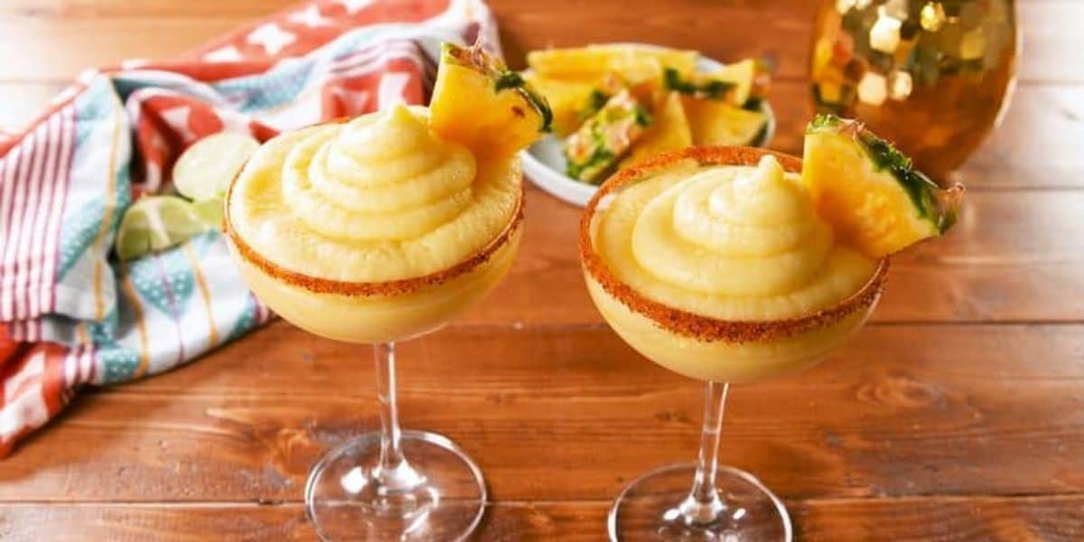 Two margarita glasses filled with a creamy yellow frozen beverage, garnished with a pineapple slice and a chili powder rim. Behind them, a colorful striped cloth napkin, lime slices, and a bowl of pineapple pieces are on a wooden table.