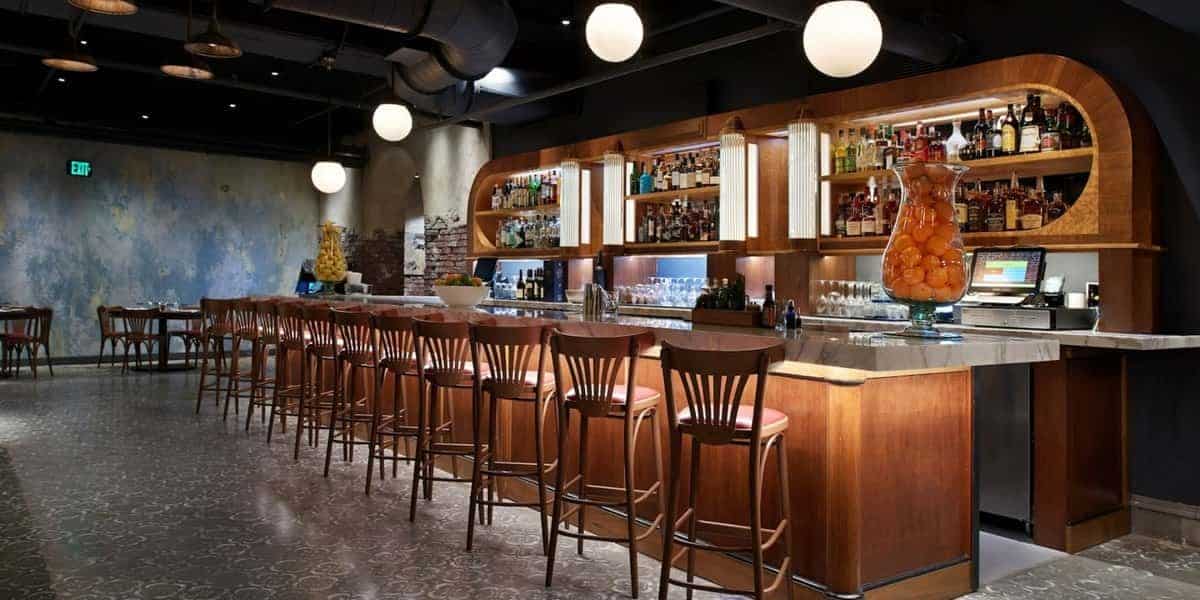A modern bar with a long wooden counter lined with high wooden chairs, illuminated by spherical pendant lights. The back wall, reminiscent of Pecos Bill's legendary hideout, is stocked with various bottles of alcohol. The bar exudes an elegant, stylish atmosphere with a few tables set at a distance.