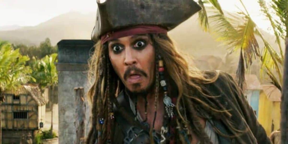 A man dressed as a pirate with a large hat, long dreadlocks, and heavy eye makeup is making a surprised or shocked facial expression. Palm trees and rustic buildings are visible in the blurred background, suggesting a tropical setting.