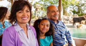 A smiling family is sitting on a Kilimanjaro Safaris ride, with a young girl nestled between two adults. They are enjoying the stunning view, while an elephant meanders in the sunlit background. The scene is bright and cheerful.
