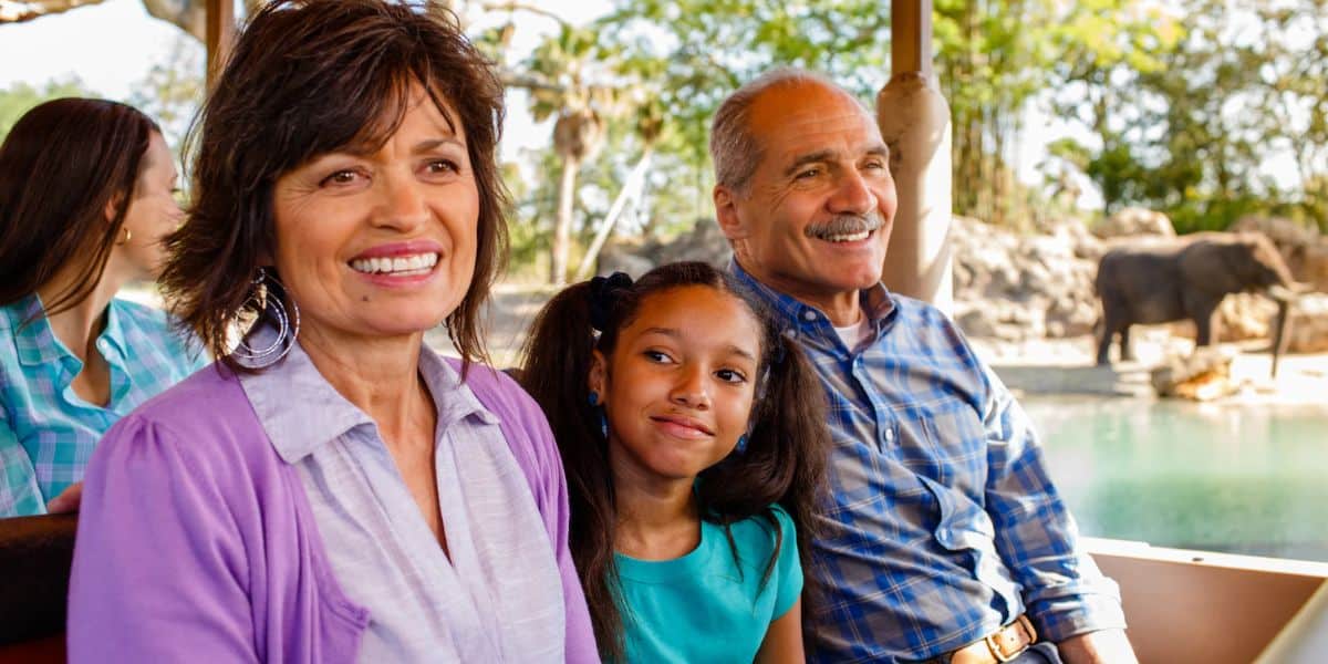 A smiling family is sitting on a Kilimanjaro Safaris ride, with a young girl nestled between two adults. They are enjoying the stunning view, while an elephant meanders in the sunlit background. The scene is bright and cheerful.