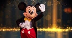 Mickey Mouse is waving and smiling while standing on a stage. He is wearing his signature outfit, which includes red shorts with white buttons, a black tuxedo jacket, a white shirt, and a yellow bow tie. Sparkling golden lights and a dark background create a festive atmosphere.