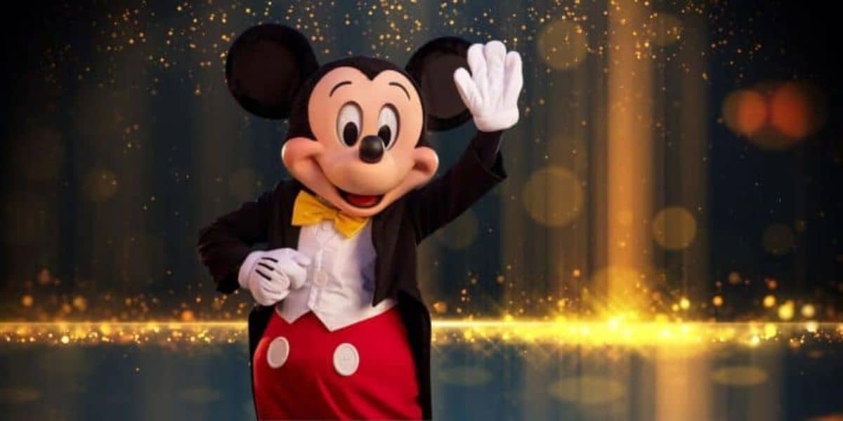 Mickey Mouse is waving and smiling while standing on a stage. He is wearing his signature outfit, which includes red shorts with white buttons, a black tuxedo jacket, a white shirt, and a yellow bow tie. Sparkling golden lights and a dark background create a festive atmosphere.