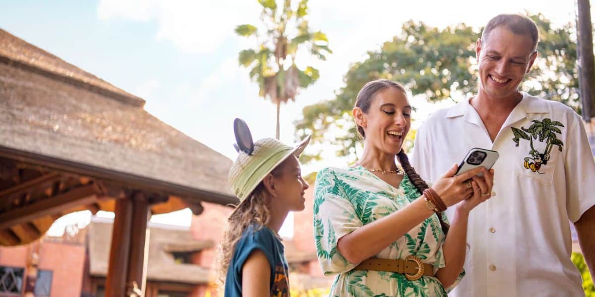 A family enjoys their time outdoors. A young child wearing a hat with mouse ears looks up at a woman holding a smartphone, who is smiling, perhaps using the chat feature on Walt Disney World's website. A man in a white shirt stands next to them, also smiling. They are near a thatched-roof building and tall palm trees.