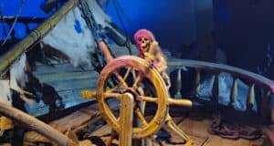 A Halloween display features a skeleton pirate dressed in tattered clothes, gripping the wheel of a decrepit ship. Reminiscent of Pirates of the Caribbean, the scene is illuminated with eerie blue and purple lighting, enhancing the spooky atmosphere.
