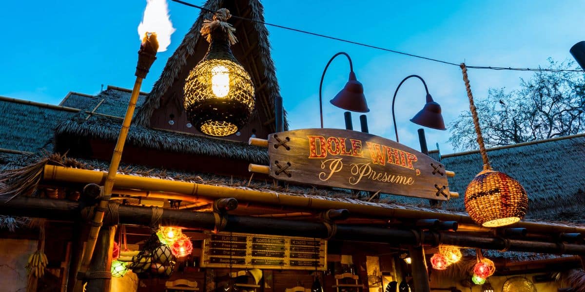 A tiki-themed outdoor restaurant with a bamboo structure, hay roofing, and colorful hanging lanterns. The wooden sign reads "Pecos Bill's Tiki Juice Bar" with a smaller sign below saying "I presume." Decorative baskets and torches illuminate the vibrant setting.
