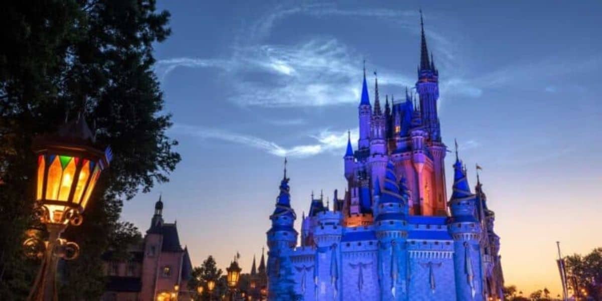 The image shows a majestic, illuminated castle at dusk, likely from a famous theme park, with its towers and spires glowing against a deep blue sky. A vintage lamppost with multicolored glass panels is visible on the left, adding to the enchanting atmosphere.