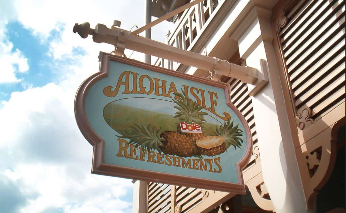A hanging sign reads "Aloha Isle Refreshments" with a Dole logo, featuring a pineapple design. Famous for its Magic Kingdom DOLE Whip, the sign is mounted on a decorative bracket attached to a building with a cloudy sky in the background.