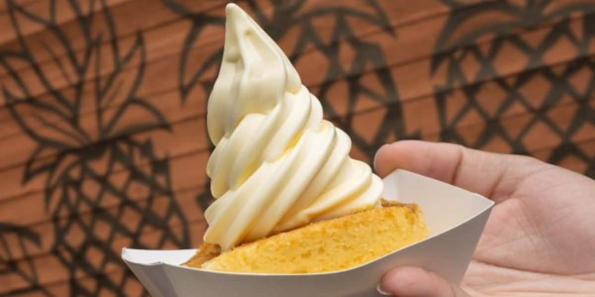 A person holds a swirl of soft-serve pineapple ice cream atop a slice of golden cake in a white paper tray. The background features a brown wall with pineapple outlines.