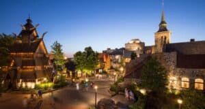 A picturesque village scene at dusk with cobblestone paths and warm lights creates a magic kingdom feel as people stroll. Wooden and stone buildings, featuring unique architectural designs like a tower and church spire, are surrounded by lush greenery, offering serene spots for water refill moments.