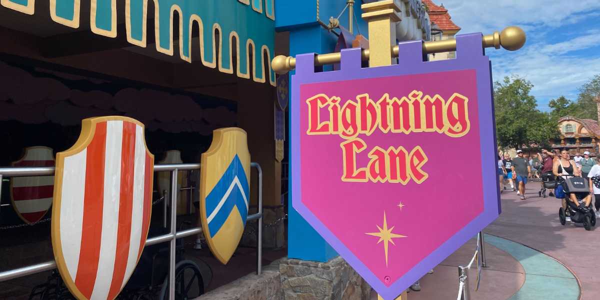 A sign for "Lightning Lane" is displayed against a colorful medieval-themed backdrop, featuring red and white striped and blue shield decorations. People walk in the background under a partly cloudy sky.