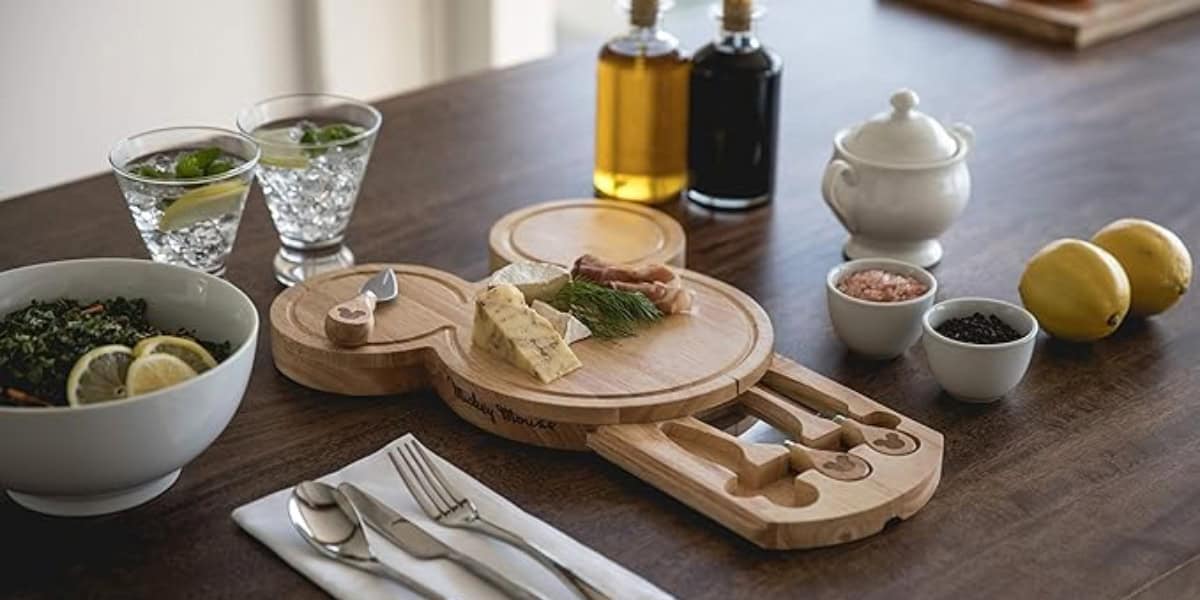 A wooden charcuterie board with assorted cheeses, cured meats, and garnishes is on a table alongside two glasses of cocktails, a bowl of salad with lemon slices, a salt dish, and utensils. Two bottles of oil and lemons, along with a sugar bowl, are in the background.