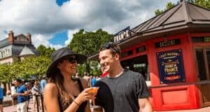 A couple smiles and holds drinks in front of a red food booth labeled "France" at an outdoor festival. The scene is lively with people walking around, and there are trees and a building in the background under a partly cloudy sky.