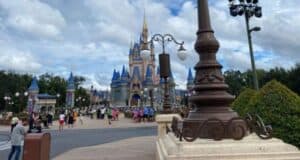 A view of a theme park with people walking in the foreground and a castle with blue and gold turrets in the background. Lamp posts and landscaped greenery line the path under a partly cloudy sky.