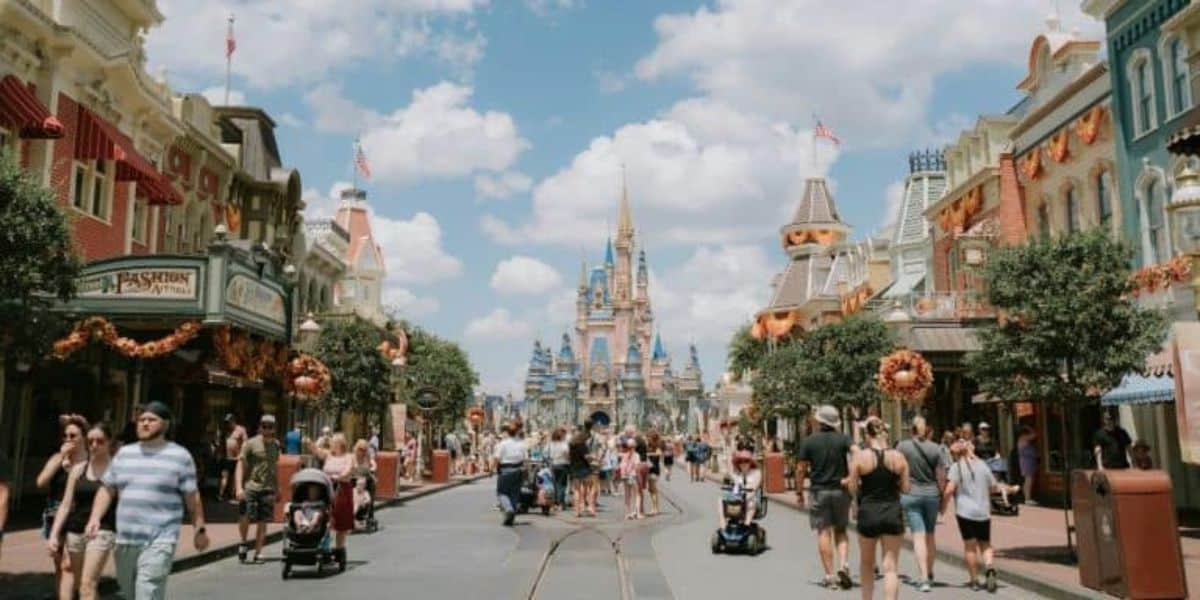 People stroll along a lively street lined with shops adorned with autumn decorations. In the background, a fairy tale castle stands under a blue sky with fluffy clouds. Families and tourists enjoy the bustling atmosphere.