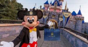 Mickey Mouse, dressed in a tuxedo and hat, stands in front of a pink and blue castle, holding a blue gift bag with a gold design. The setting is a theme park with decorative flags adorning the castle.