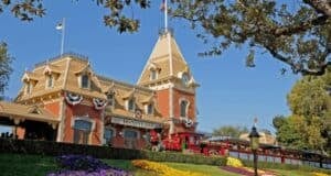 A charming red train is parked at a station surrounded by lush greenery and colorful flowers. The building features a clock tower and is adorned with flags and decorative elements under a clear blue sky.