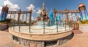 A wide-angle view of a fairytale castle with towers and spires, surrounded by ornate wrought iron fencing. The foreground shows a circular paved area. The sky is clear with a few clouds, creating a picturesque scene.