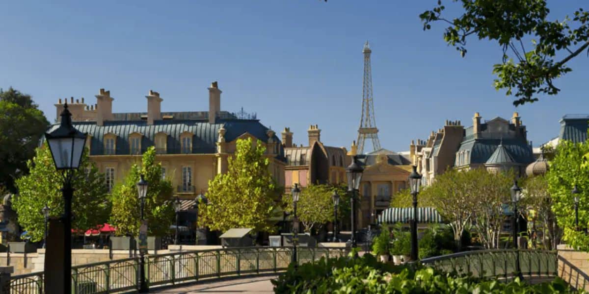 Immerse yourself in a picturesque European-style village, reminiscent of Epcot's charm, with pastel-colored buildings and grey rooftops. The Eiffel Tower graces the clear blue sky, while lush green trees and vintage street lamps complete this enchanting scene.
