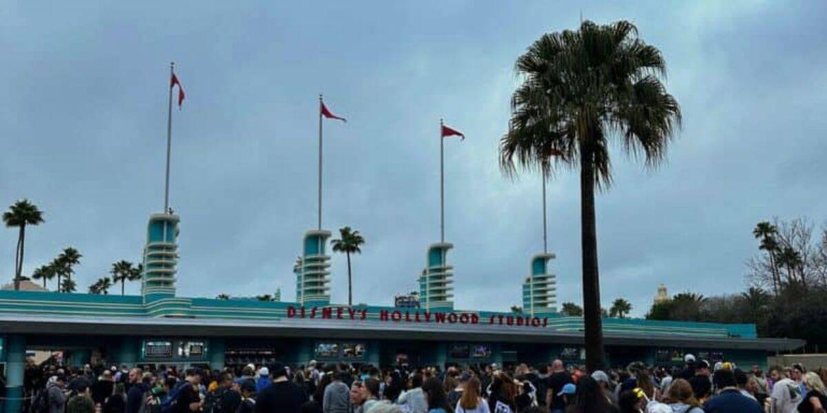 Crowd gathered outside the entrance of Disney's Hollywood Studios, with art deco architecture and palm trees. Three flags are flying, and the sky is overcast.