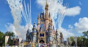 A grand castle stands against a blue sky, surrounded by white clouds. Colorful fireworks burst in the background. In front of the castle, costumed characters gather on the stage, with tall green trees visible on the sides.