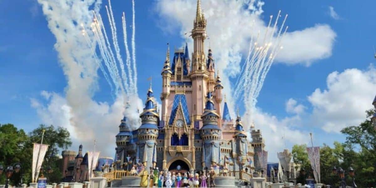 A grand castle stands against a blue sky, surrounded by white clouds. Colorful fireworks burst in the background. In front of the castle, costumed characters gather on the stage, with tall green trees visible on the sides.