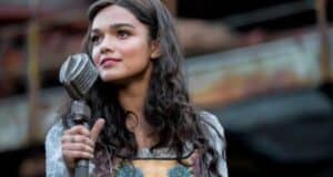 A woman with long, wavy hair stands outdoors holding a vintage microphone. She is wearing a dress with floral embroidery and appears to be singing or speaking, with a thoughtful expression. The background is blurred, showing a rustic setting.
