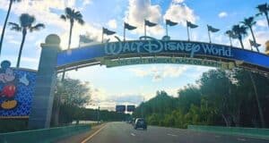 Entrance sign to Walt Disney World with the text "The Most Magical Place on Earth" displayed prominently. A Mickey Mouse image is on the left side of the sign. The road leads past the entrance with trees on both sides and blue sky with clouds overhead.
