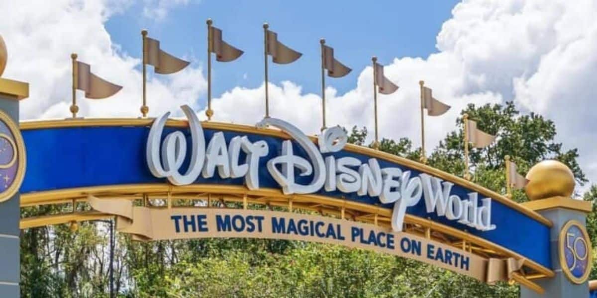 Entrance sign to Walt Disney World with a blue and gold design, featuring the phrase "The Most Magical Place on Earth." The sky is partly cloudy, and trees are visible in the background.