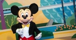 A person in a Mickey Mouse costume stands smiling in front of a colorful background featuring geometric shapes and a monorail, embodying the spirit of Character Sightings at EPCOT. Mickey is dressed in red shorts, a yellow bow tie, and a black jacket.