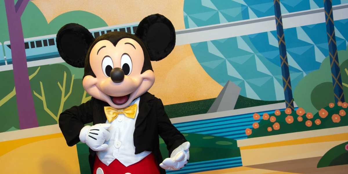 A person in a Mickey Mouse costume stands smiling in front of a colorful background featuring geometric shapes and a monorail, embodying the spirit of Character Sightings at EPCOT. Mickey is dressed in red shorts, a yellow bow tie, and a black jacket.